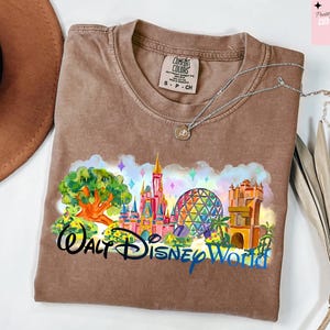 K&ouml;nnte beinhalten: Ein braunes Comfort Colors T-Shirt mit einer farbenfrohen Grafik von Walt Disney World. Das Design umfasst ein Schloss, Spaceship Earth und den Text "Walt Disney World". Eine silberne Halskette liegt auf dem Shirt.
