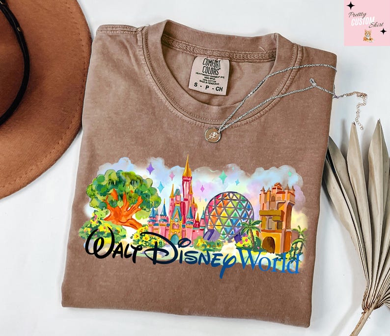 K&ouml;nnte beinhalten: Ein braunes Comfort Colors T-Shirt mit einer farbenfrohen Grafik von Walt Disney World. Das Design umfasst ein Schloss, Spaceship Earth und den Text "Walt Disney World". Eine silberne Halskette liegt auf dem Shirt.
