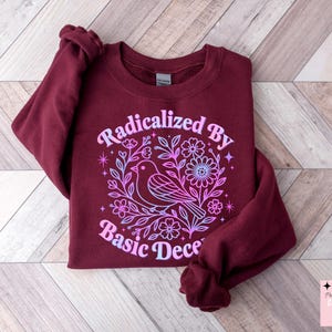 Peut inclure: Sweat-shirt bordeaux avec l'inscription "Radicalized By Basic Decency" dans un motif floral avec un oiseau. Le motif présente un dégradé rose et bleu. Le sweat-shirt est de style col rond.