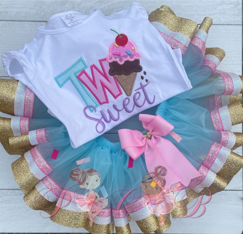 May include: A white onesie with the words "Two Sweet" and an ice cream cone graphic. The onesie is paired with a blue tulle tutu with pink and gold ribbon trim.