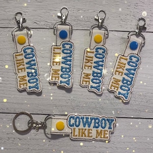 May include: Five white keychains with blue and gold embroidery that reads "Cowboy Like Me". Each keychain has a small blue or yellow circle on the front.