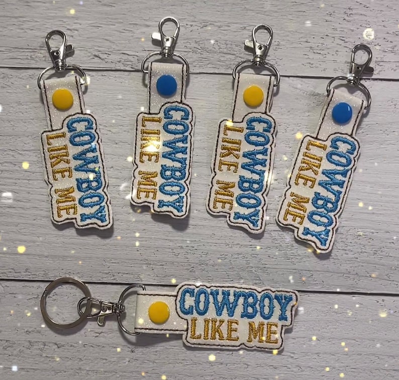 May include: Five white keychains with blue and gold embroidery that reads "Cowboy Like Me". Each keychain has a small blue or yellow circle on the front.