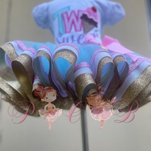 May include: A light blue tulle tutu with gold and pink glitter ribbon trim. Two ballerina figures are attached to the tutu. The tutu is on a white dress form with a white short-sleeved shirt that says "Two Cute" in pink, blue, and green letters.