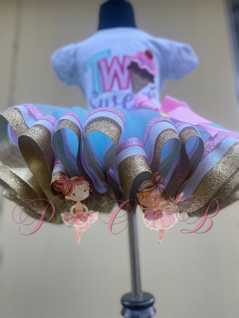 May include: A light blue tulle tutu with gold and pink glitter ribbon trim. Two ballerina figures are attached to the tutu. The tutu is on a white dress form with a white short-sleeved shirt that says "Two Cute" in pink, blue, and green letters.