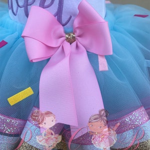 May include: A light blue tulle tutu skirt with a pink bow and gold glitter trim. The skirt has a white top with the words "So Sweet" embroidered on it. The skirt is decorated with two images of ballerinas.