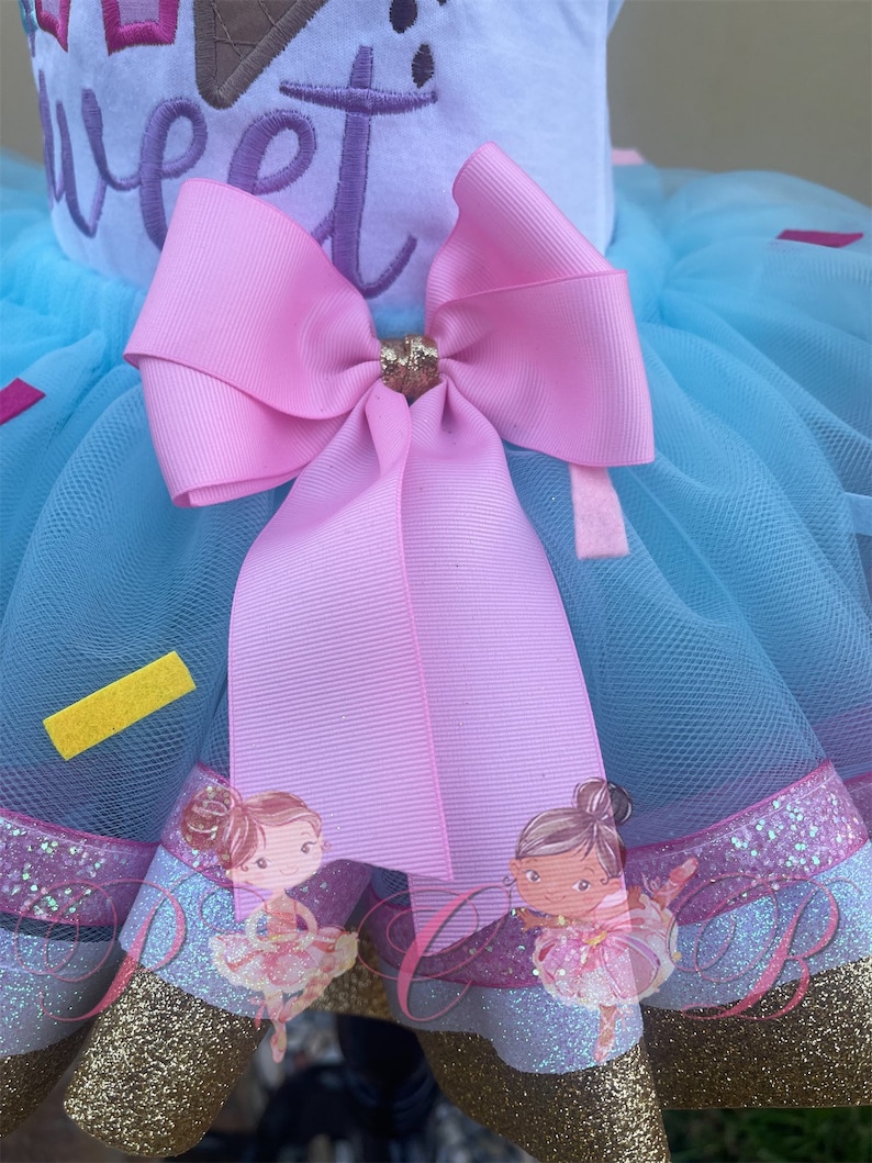 May include: A light blue tulle tutu skirt with a pink bow and gold glitter trim. The skirt has a white top with the words "So Sweet" embroidered on it. The skirt is decorated with two images of ballerinas.