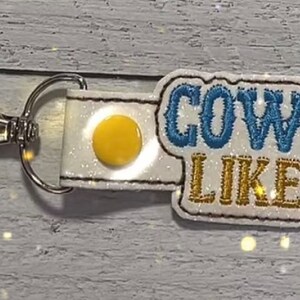 May include: White keychain with a blue and gold glitter design that says "COWBOY LIKE ME".