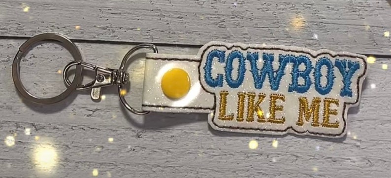 May include: White keychain with a blue and gold glitter design that says "COWBOY LIKE ME".