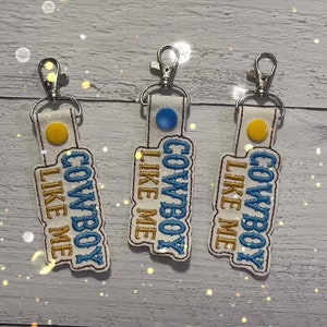 May include: Three white keychains with blue and gold embroidery that reads "Cowboy Like Me".