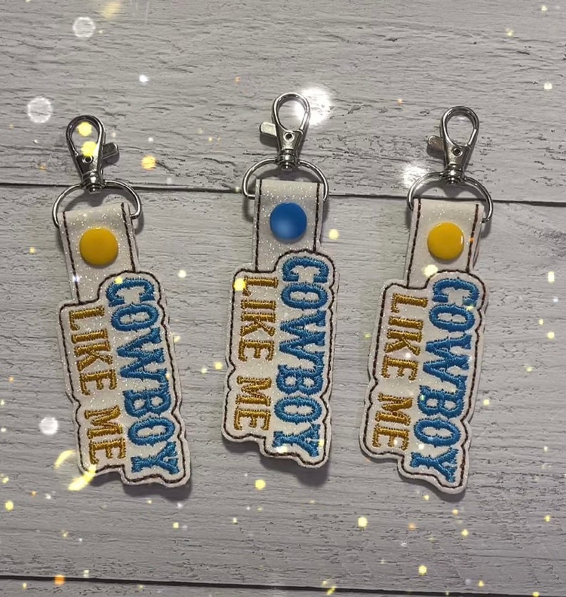 May include: Three white keychains with blue and gold embroidery that reads "Cowboy Like Me".