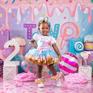 May include: A young girl wearing a white shirt with the text "Kailtyn is Two Sweet" and a blue and gold tutu stands in front of a pink and blue candy-themed backdrop. She is surrounded by colourful decorations, including a large number "2", a doughnut, and a cupcake.