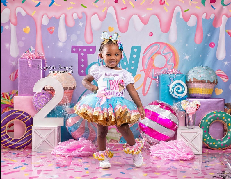 May include: A young girl wearing a white shirt with the text "Kailtyn is Two Sweet" and a blue and gold tutu stands in front of a pink and blue candy-themed backdrop. She is surrounded by colourful decorations, including a large number "2", a doughnut, and a cupcake.