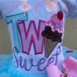 May include: A light blue tutu with a white shirt that says "Two Sweet" in colourful embroidered letters. The shirt also features two ballerina figures and an ice cream cone with sprinkles. A pink bow is attached to the side of the tutu.