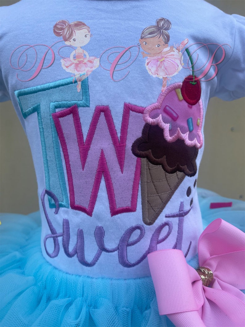 May include: A light blue tutu with a white shirt that says "Two Sweet" in colourful embroidered letters. The shirt also features two ballerina figures and an ice cream cone with sprinkles. A pink bow is attached to the side of the tutu.