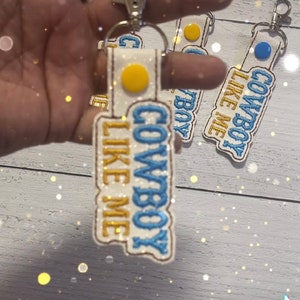 May include: A white fabric keychain with blue and gold embroidery that reads "Cowboy Like Me". The keychain has a yellow circle with a white center and a blue circle with a white center.
