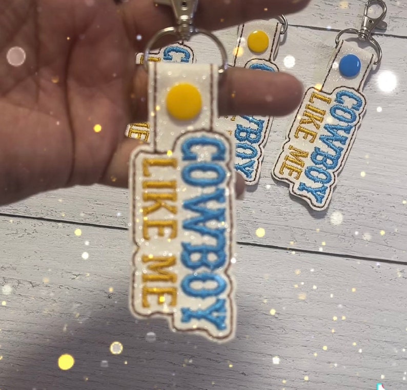 May include: A white fabric keychain with blue and gold embroidery that reads "Cowboy Like Me". The keychain has a yellow circle with a white center and a blue circle with a white center.