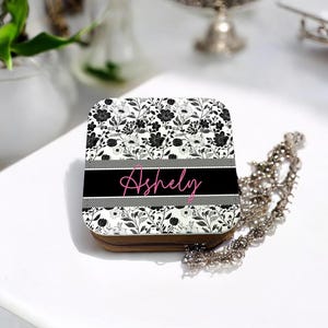 May include: A black and white floral patterned jewelry box with a zipper closure. The box has a black band with the name "Ashly" written in pink.