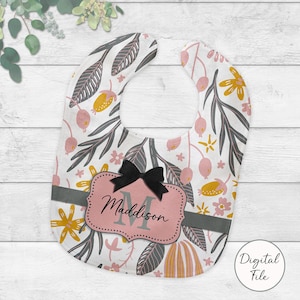 May include: A baby bib with a floral pattern in pink and grey. It has a black bow and a pink label with the name "Maddison" and the letter "M". The bib's background is white, with yellow and pink floral elements.