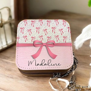 Può includere: Una scatola per gioielli rosa e bianca con un fiocco rosa e il nome "Madaline" stampato sul davanti. La scatola è decorata con un motivo di fiocchi rosa su sfondo bianco.