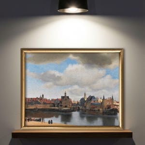 以下が含まれることがあります： 川、ボート、建物が描かれたヨーロッパの町の詳細な絵画が特徴の、額入りのジグソーパズル。 パズルは金色のフレームで、木製の棚に飾られています。 空は青く、ふわふわの白い雲があります。