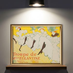 以下が含まれることがあります： 黄色い背景に「Troupe de Mlle Eglantine」の文字が書かれた、額縁に入ったヴィンテージポスター。白いドレスと帽子を身につけ、黒いストッキングと靴を履いた3人の女性が描かれています。フレームは金色です。