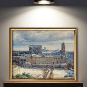 May include: A framed landscape painting depicting a snow-covered townscape with buildings and a church tower. The artwork is set in a gold-colored frame and is displayed on a wooden shelf.