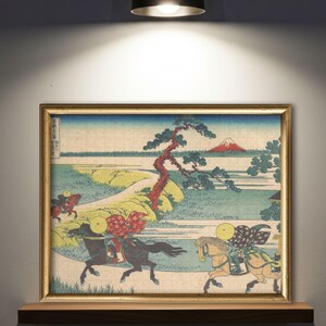 May include: Framed jigsaw puzzle featuring a Japanese landscape with riders on horseback. The artwork depicts a mountain, a winding path, and a large tree. The puzzle is in a gold-colored frame and rests on a wooden shelf.