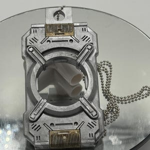 Puede incluir: Un colgante rectangular plateado con una abertura circular y dos tubos blancos. El colgante tiene un diseño industrial detallado con detalles dorados y una cadena de bolas. Probablemente sea una joya o un objeto decorativo.