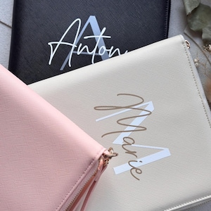 Könnte beinhalten: Drei Reißverschluss-Clutch-Geldbörsen in Schwarz, Creme und Rosa. Jede Geldbörse hat einen personalisierten Namen in Schreibschrift mit einem Großbuchstaben. Die schwarze Geldbörse hat den Namen "Anton" in Weiß, die cremefarbene Geldbörse den Namen "Marie" in Braun.