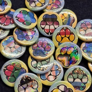 May include: A collection of colourful paw print buttons featuring various pride flag designs. The buttons are round and have a shiny, iridescent finish.