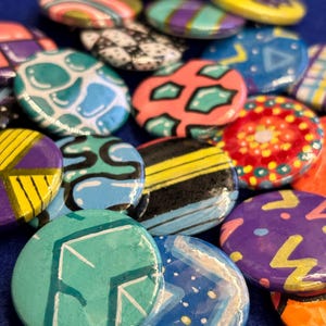 May include: A collection of colourful, hand-painted pinback buttons with various geometric and abstract designs. The buttons are made of metal and have a shiny finish.