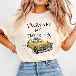 May include: A cream-colored short-sleeve t-shirt featuring the black text I SURVIVED MY TRIP TO NYC above a graphic illustration of a yellow taxi cab with a simple stick figure driver and a checkered stripe along the side of the vehicle.