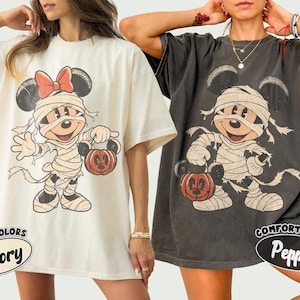 May include: Two oversized t-shirts in ivory and pepper colors, each featuring a graphic print of a cartoon mouse wrapped in bandages like a mummy, wearing a bow and holding a pumpkin bucket. The text Comfort Colors Ivory and Pepper is visible.