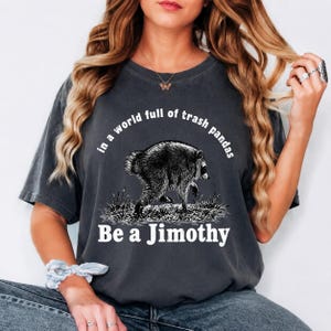 May include: A charcoal gray short-sleeve t-shirt featuring a black and white illustration of a raccoon walking through grass. The text reads, "in a world full of trash pandas Be a Jimothy" in white lettering across the front of the garment.