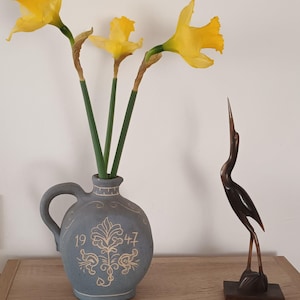 Può includere: Una natura morta con tre narcisi gialli in un vaso di ceramica grigia con manico e una scultura di airone in legno marrone scuro. Il vaso presenta un motivo decorativo e i numeri "1947". I fiori hanno steli verdi.