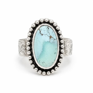 May include: An oval-shaped turquoise gemstone ring set in sterling silver. The central light blue stone features dark brown matrix veining and is surrounded by a beaded silver border. The ring band has an etched floral pattern on the sides.