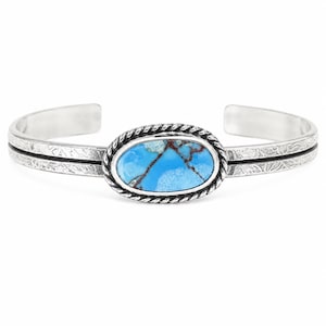 May include: A silver cuff bracelet featuring a central oval turquoise stone with brown matrix veining. The stone is set within a twisted rope border. The band has a textured, etched surface with a horizontal line running through the center.