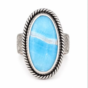 May include: An oval ring featuring a bright blue stone with a white horizontal vein, set in a silver bezel with a twisted rope border. The silver band displays an intricate engraved pattern on the sides, providing a textured finish.