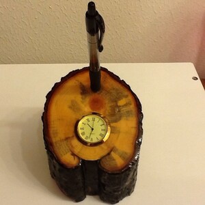 May include: A wooden pen holder with a clock embedded in the top. The holder is made from a tree trunk slice with a dark brown bark and a light brown interior. A black pen is inserted into the top of the holder.