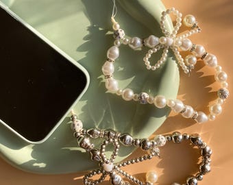 May include: Two phone charms with pearl and silver beads. One charm features a pearl bow with a silver accent. The other charm has a silver bow with a pearl accent.