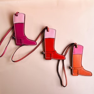 May include: Three colorful leather boot keychains. Each boot is a different color: fuchsia, red, and orange, with a pink top and brown stitching. Each keychain has a matching leather strap and a silver-tone metal clasp.