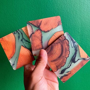 May include: Three square coasters with a distinctive design. They combine natural wood grain with a light green, translucent resin. The wood displays a warm, reddish-brown colour, while the resin has dark, swirling patterns. Each coaster is approximately 10 cm x 10 cm.