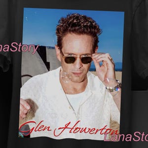 Puede incluir: Camiseta unisex negra con una foto de un hombre con gafas de sol. La imagen de la camiseta tiene un fondo de cielo azul. El texto "Glen Howerton" está impreso en rojo. La camiseta también tiene el texto "UNISEX T-SHIRT".