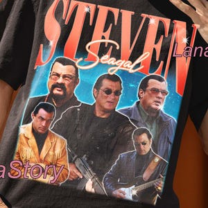 May include: Black unisex t-shirt featuring a graphic of Steven Seagal with the text "STEVEN Seagal". The shirt has a crew neck and short sleeves. The shirt also has the text "UNISEX T-SHIRT" at the top.