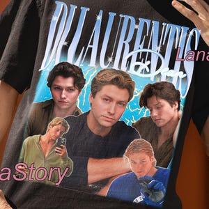 May include: Black unisex t-shirt featuring a graphic design with the text "DI LAURENTIS" in blue and white. The design includes images of several men. The words "UNISEX T-SHIRT" are at the top.