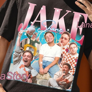 May include: Black unisex t-shirt with a graphic featuring the name "Jake" in pink and white, and multiple images of a person. The shirt also has the text "Shane".