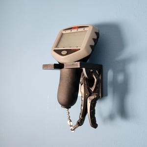 May include: A black and gray digital scale with a chain hanging from the bottom. The scale is mounted on a black wall mount with a black and gray clamp.
