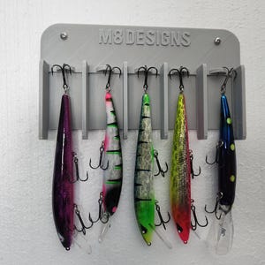 May include: A gray wall-mounted fishing lure rack with the text "M8 DESIGNS" printed on it. Six fishing lures are hanging on the rack. The lures are in various colors, including purple, green, yellow, and blue.