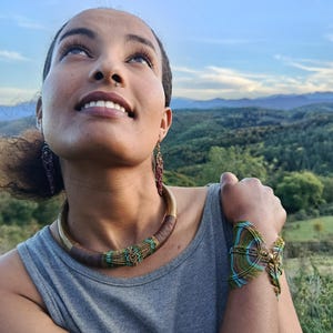 May include: A woman wearing a handmade jewelry set. The set includes a gold-toned choker necklace with brown and teal accents, matching earrings, and a bracelet. The jewelry is set against a backdrop of a green landscape and blue sky.