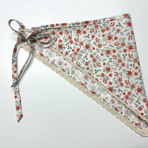 May include: A white triangular bandana with a floral pattern of red flowers and green leaves. The bandana has a cream-colored lace trim along the edges and a tie closure. The fabric is soft and lightweight.